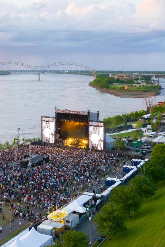 An aerial view of RiverBeat Music Fest, with a large crowd gathered in front of a large stage. The Mississippi River, Hernando de Soto Bridge and Memphis skyline are in the background.