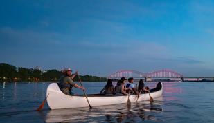 A group of people row a multi-person canoe on the Mississippi River with an illuminated bridge in the background.